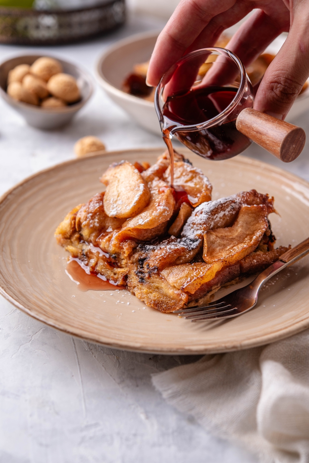 A serving of French toast covered in stewed apples and powdered sugar and a bowl of maple syrup is being poured over the French toast.