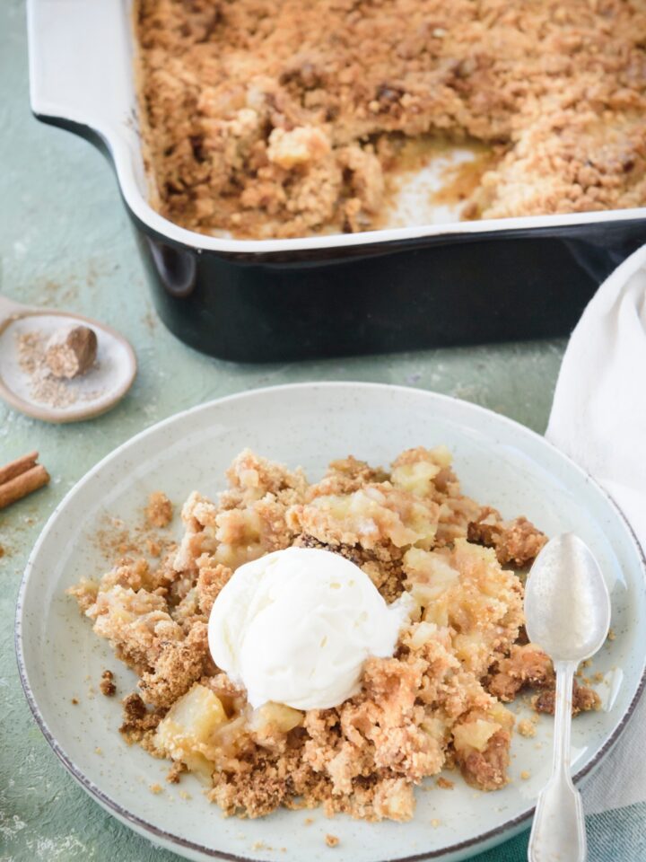 Apple crumble on a plate with a scoop of vanilla ice cream on top and a fork is also on the plate. In the background is a baking dish filled with more crumble.