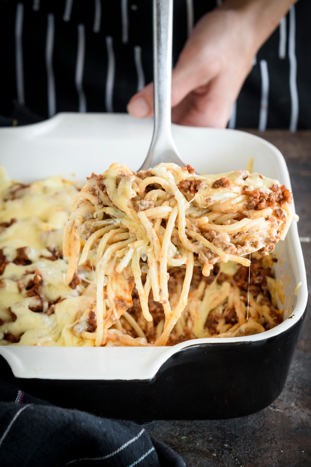 A serving spoon holding a scoop of spaghetti noodles covered in a creamy red sauce with chunks of ground beef and strings of melted cheese.