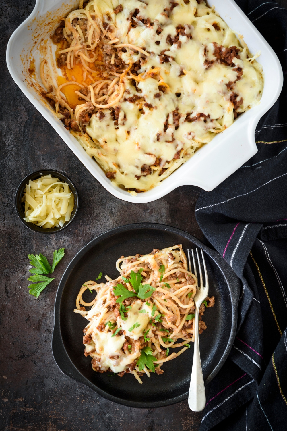 A bowl of spaghetti topped with fresh herbs and melted cheese next to a casserole dish filled with more spaghetti.