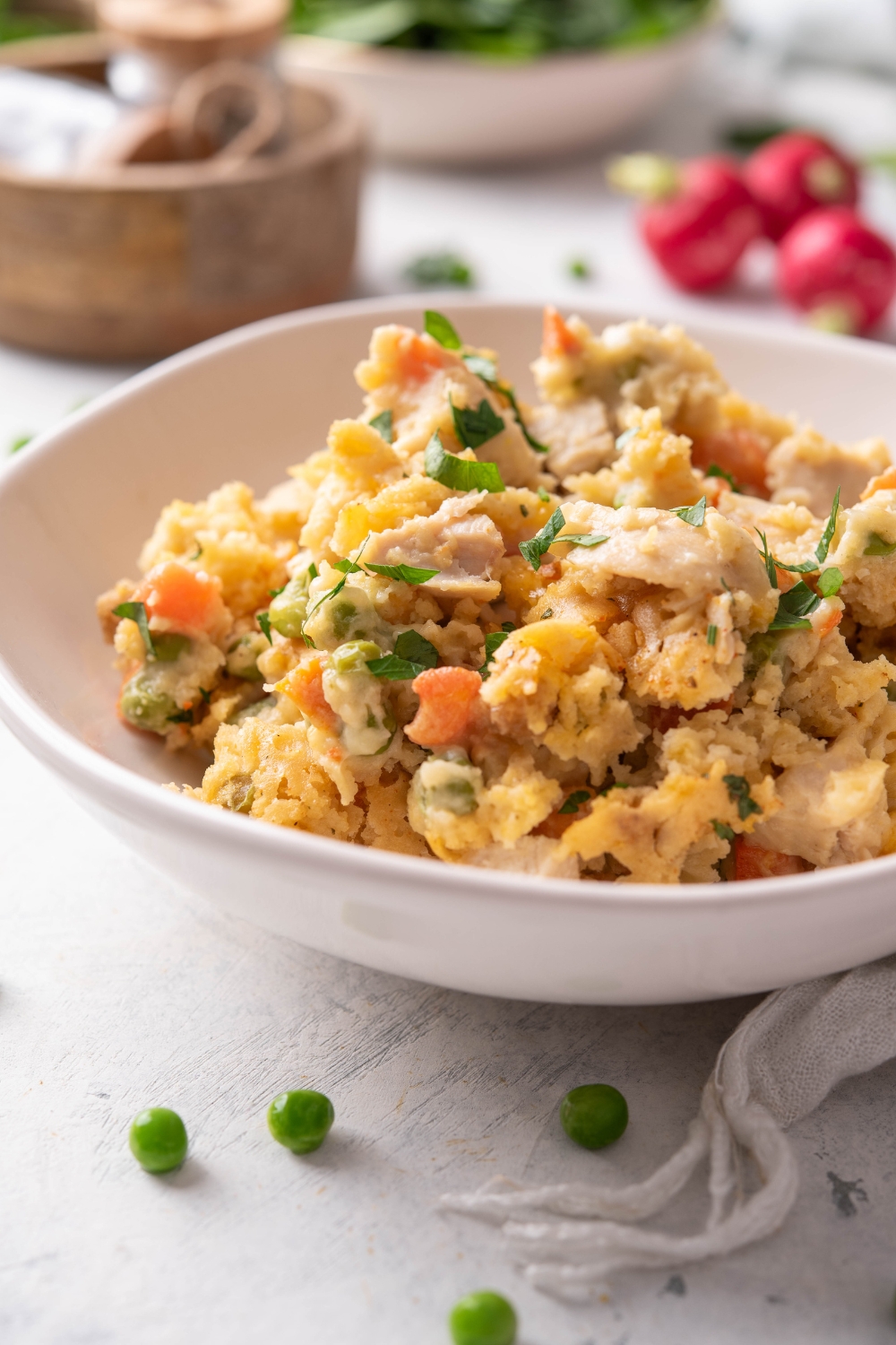 A bowl of chicken casserole with cooked chicken in a creamy sauce mixed with carrots, peas, and a crumbly biscuit topping and garnished with fresh herbs.