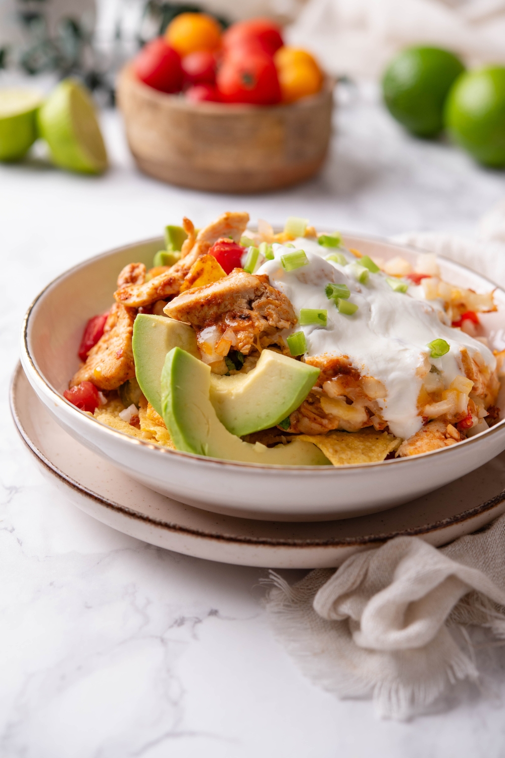 A bowl of chicken nachos covered in queso sauce, garnished with pico de gallo and green onions, with sliced avocado on the side.