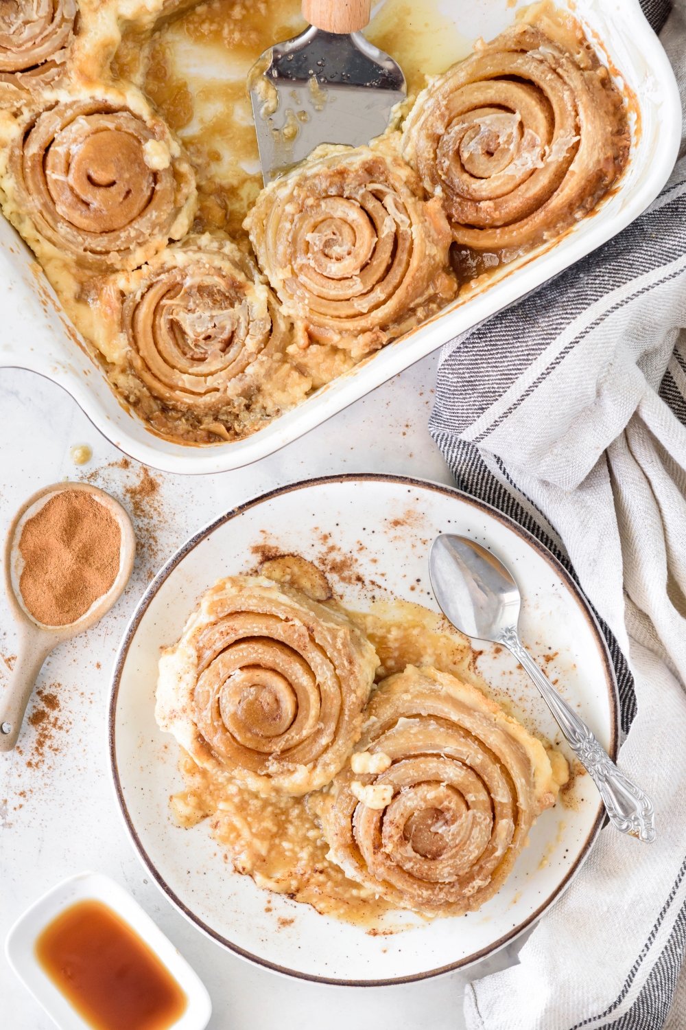 Two cinnamon rolls on a plate dusted with cinnamon. The rest of the cinnamon rolls are in a baking dish next to the plate and a third cinnamon roll is being scooped out of the baking dish.