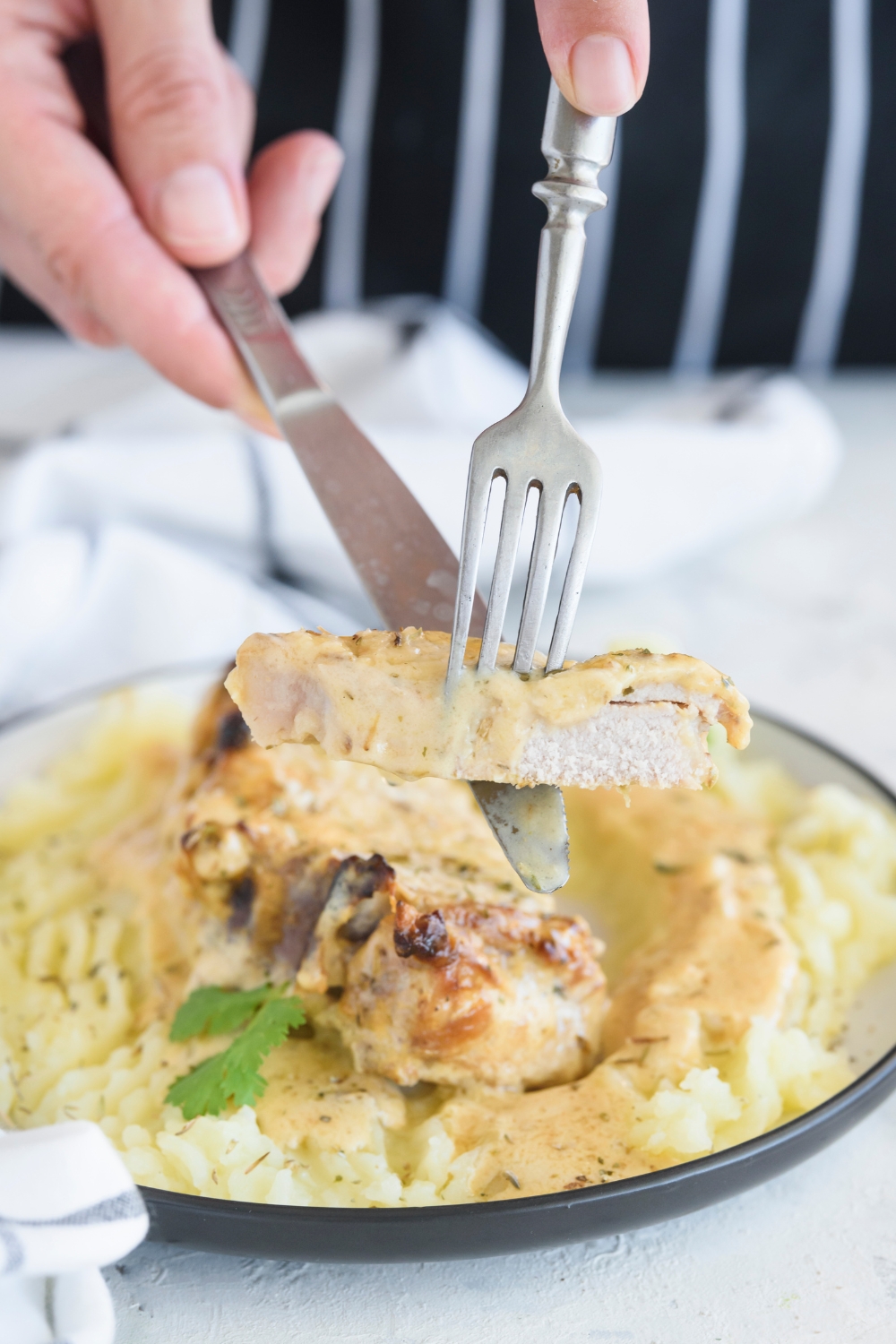 A person using a fork and knife to hold up a piece of pork chop covered in a brown cream sauce. The rest of the pork chop is in the background.