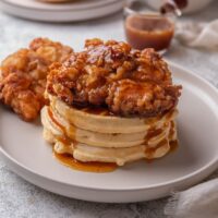 A stack of three waffles with a piece of fried chicken on top covered in a glaze that is dripping down the waffles. There is a second piece of fried chicken next to the stack of waffles.
