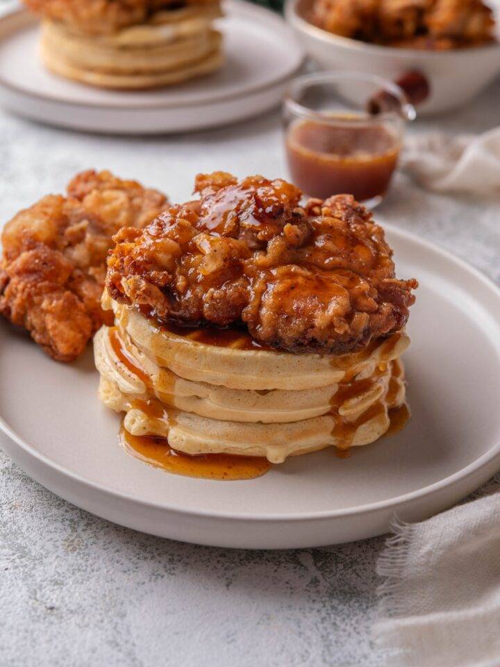 A stack of three waffles with a piece of fried chicken on top covered in a glaze that is dripping down the waffles. There is a second piece of fried chicken next to the stack of waffles.