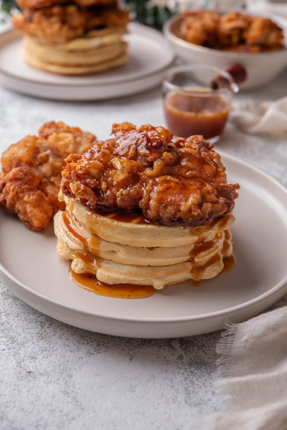 A stack of three waffles with a piece of fried chicken on top covered in a glaze that is dripping down the waffles. There is a second piece of fried chicken next to the stack of waffles.