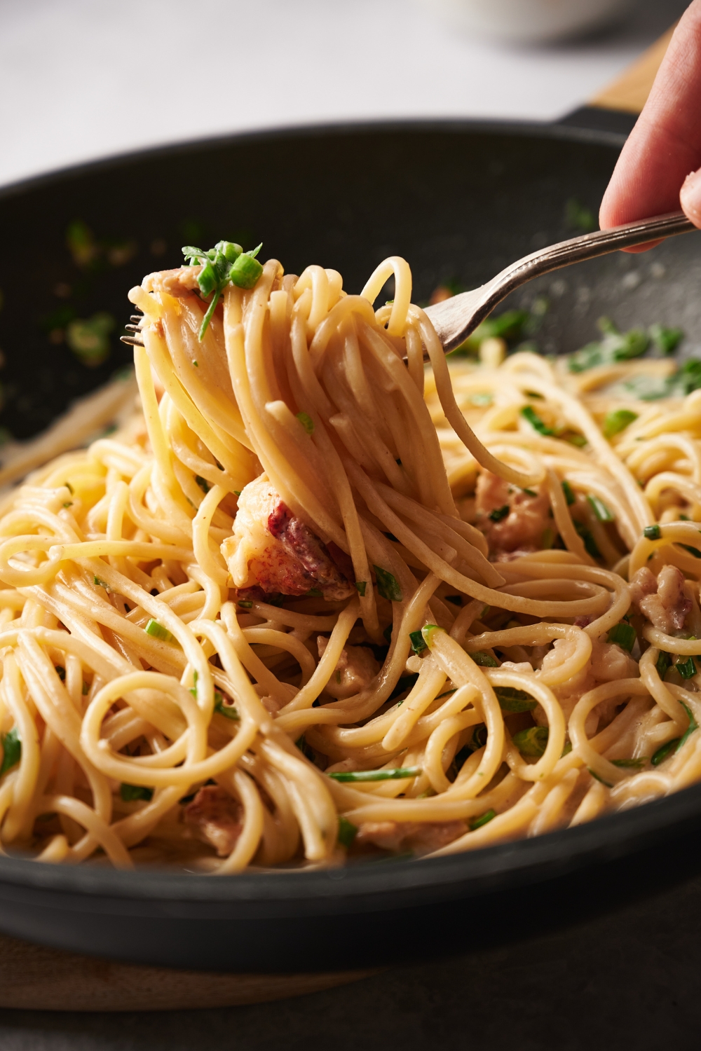 A fork taking a scoop of cooked spaghetti from a bowl. The spaghetti has lobster meat and green onions mixed in it.