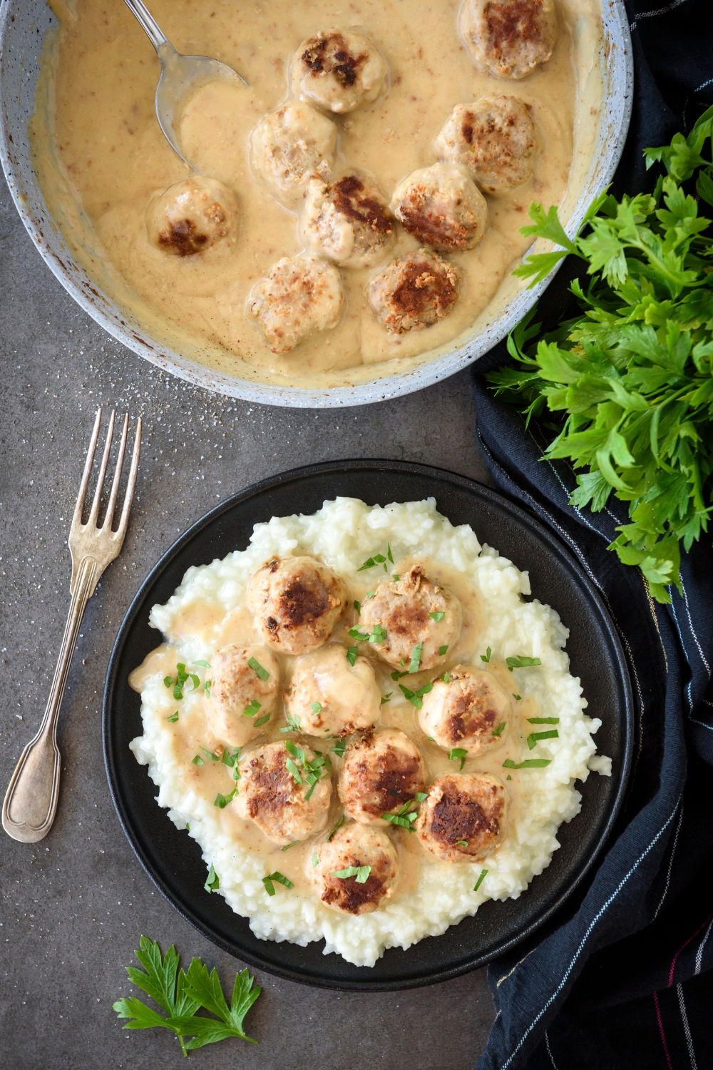 A plate of meatballs covered in fresh herbs and a creamy gravy on a bed of white rice. The rest of the meatballs and sauce are in a skillet next to the plate.