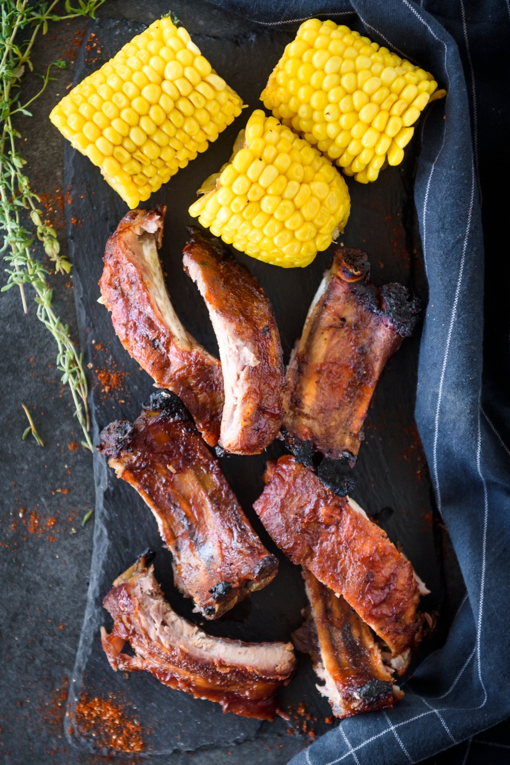 A pile of ribs covered in BBQ sauce on a black platter with three sliced corn cobs.