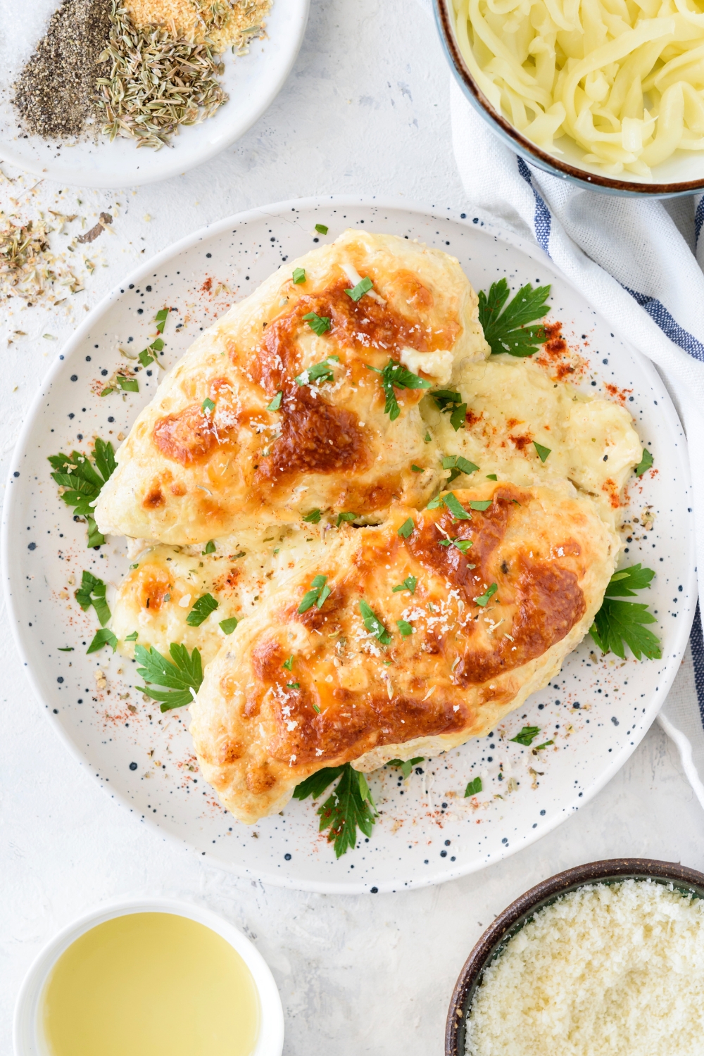 Two freshly baked chicken breasts on a plate garnished with fresh herbs, salt, and spices. The chicken is covered in a creamy sauce.