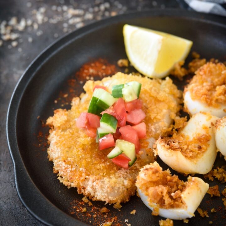 A breaded calamari steak topped with cucumber and diced tomato on a plate with a lemon wedge and chunks of sliced calamari.