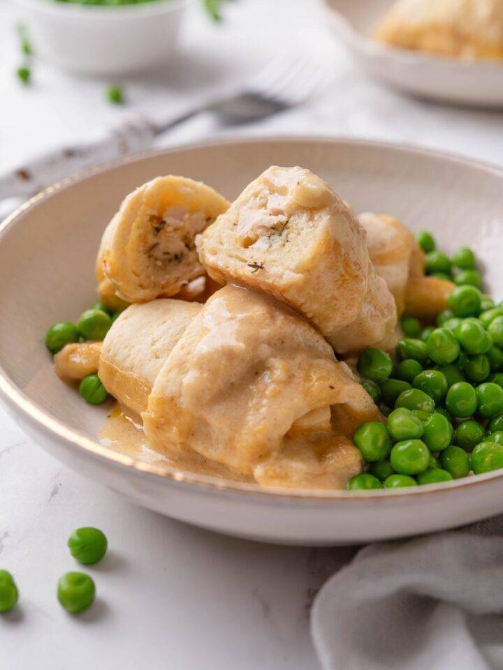 A bowl with three stuffed crescent rolls covered in cream sauce and one crescent roll has been cut in half to reveal the chicken filling. The crescent rolls are served with a side of green peas.