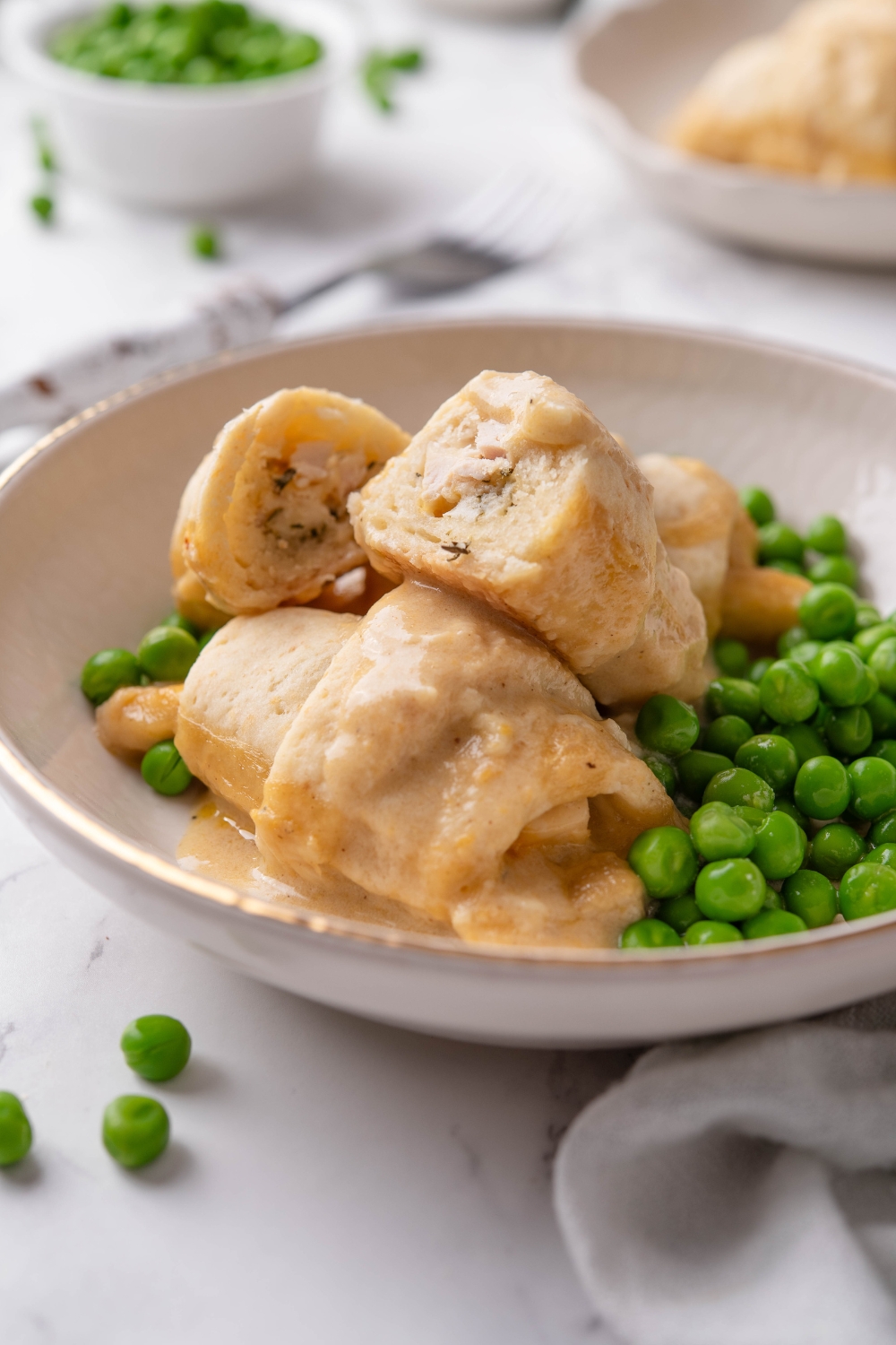 A bowl with three stuffed crescent rolls covered in cream sauce and one crescent roll has been cut in half to reveal the chicken filling. The crescent rolls are served with a side of green peas.