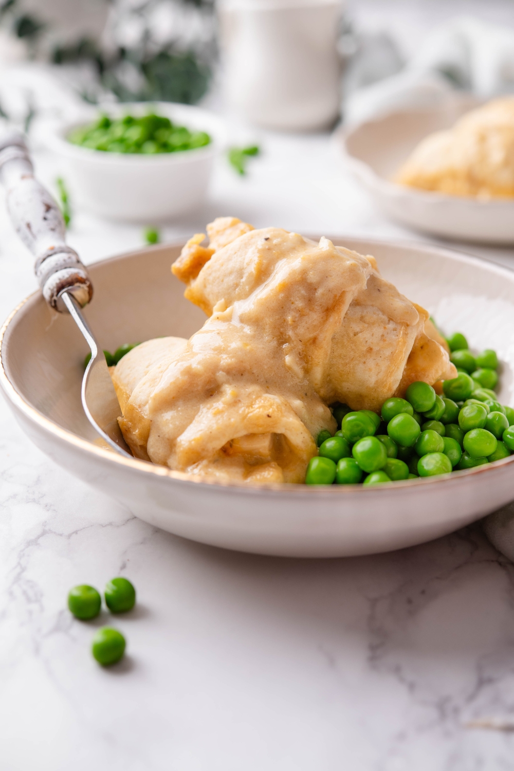 A bowl with two stuffed crescent rolls covered in cream sauce next to a pile of cooked green peas and a fork is in the bowl.