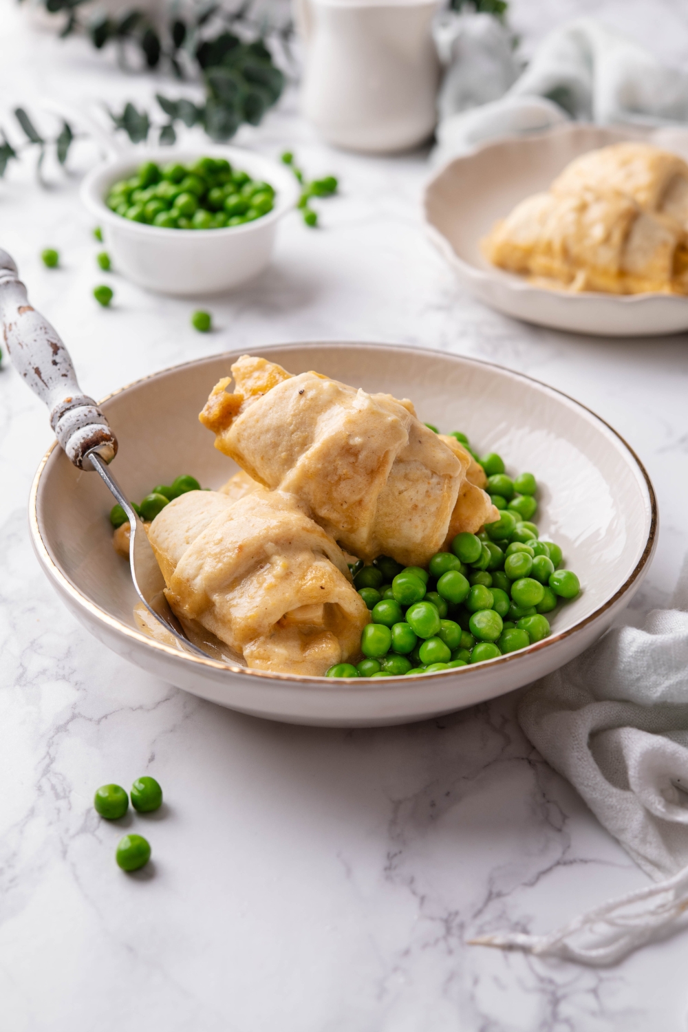 A bowl with two stuffed crescent rolls covered in cream sauce next to a pile of cooked green peas and a fork is in the bowl.