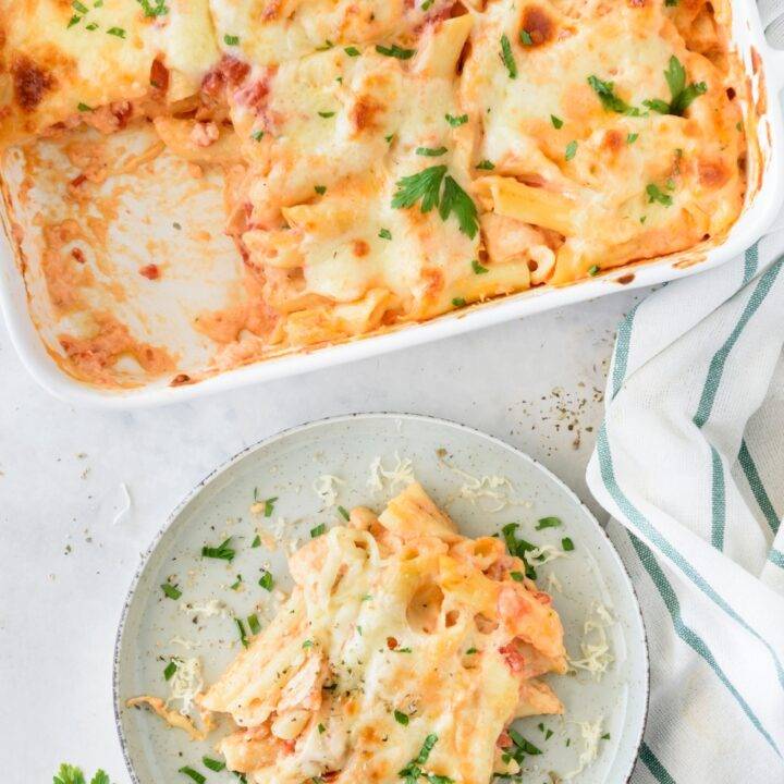 A plate of chicken pasta bake covered in melted cheese and fresh herbs next to a baking dish filled with the rest of the pasta.