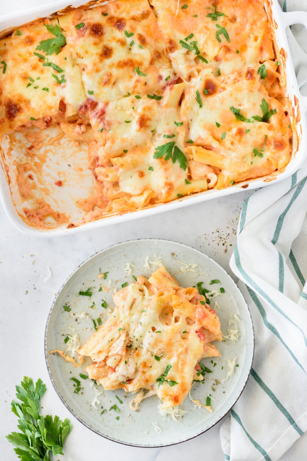 A plate of chicken pasta bake covered in melted cheese and fresh herbs next to a baking dish filled with the rest of the pasta.