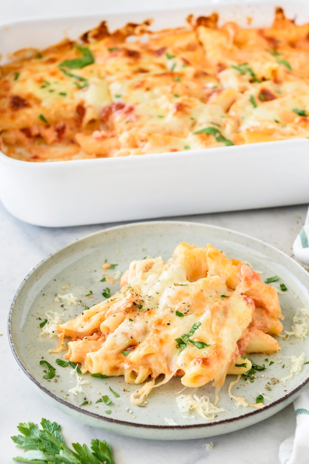 A plate of chicken pasta bake covered in melted cheese and fresh green herbs. The rest of the pasta is in a baking dish in the background.