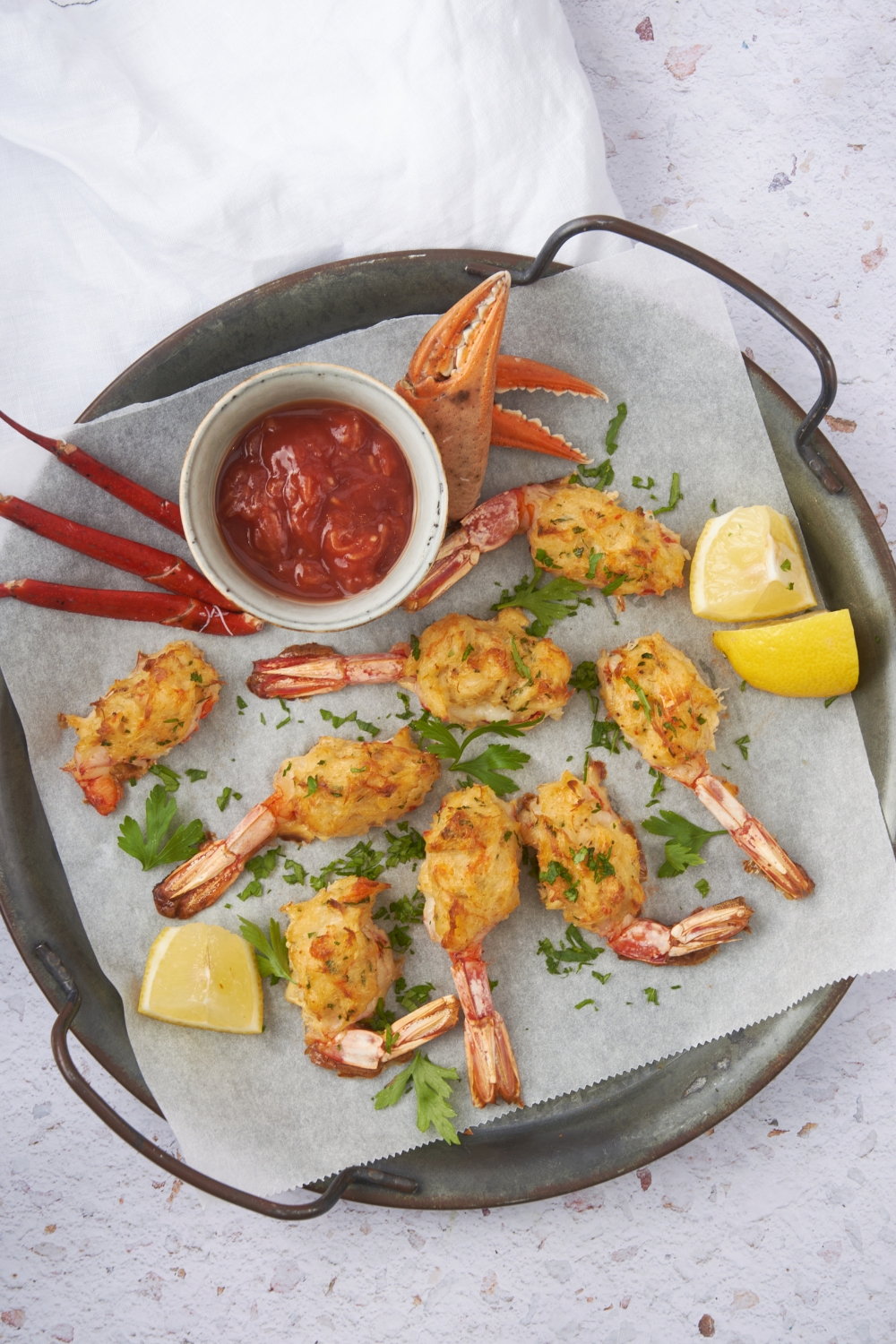 A pan lined with parchment paper filled with baked shrimp stuffed with crab meat. Surrounded the shrimp are lemon wedges, fresh herbs, a bowl of cocktail sauce, and a crab claw.