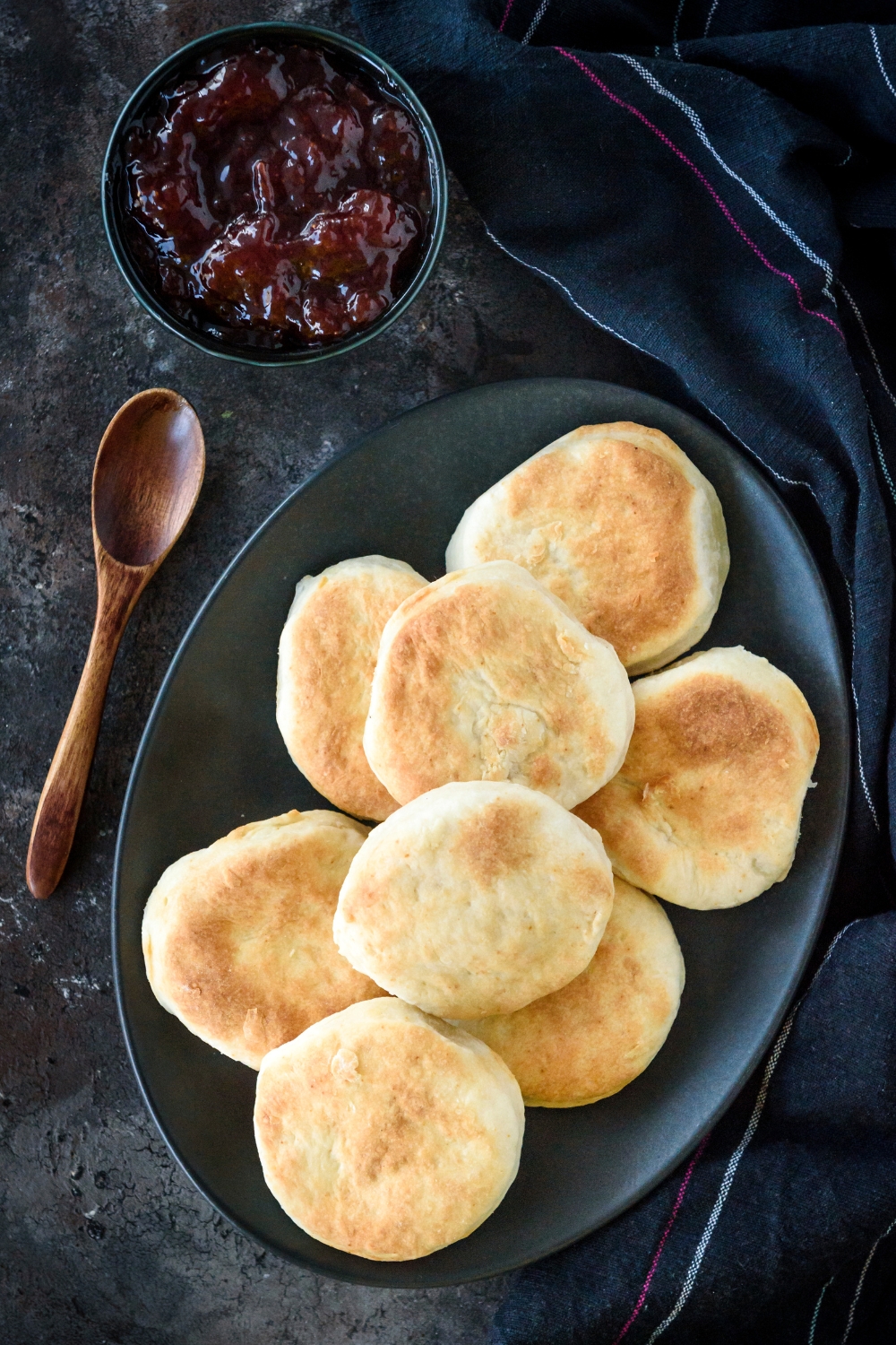 A pile of biscuits on a black plate next to a spoon and a bowl of jam.