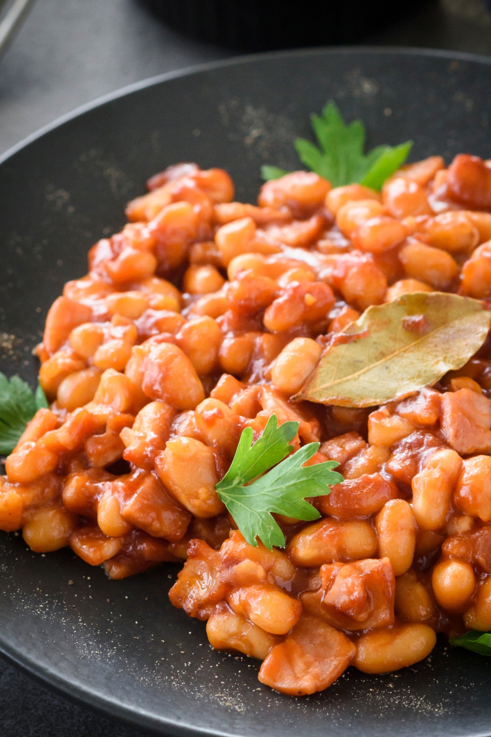 A completed dish of baked beans on a black plate with garnish greens and a bay leaf
