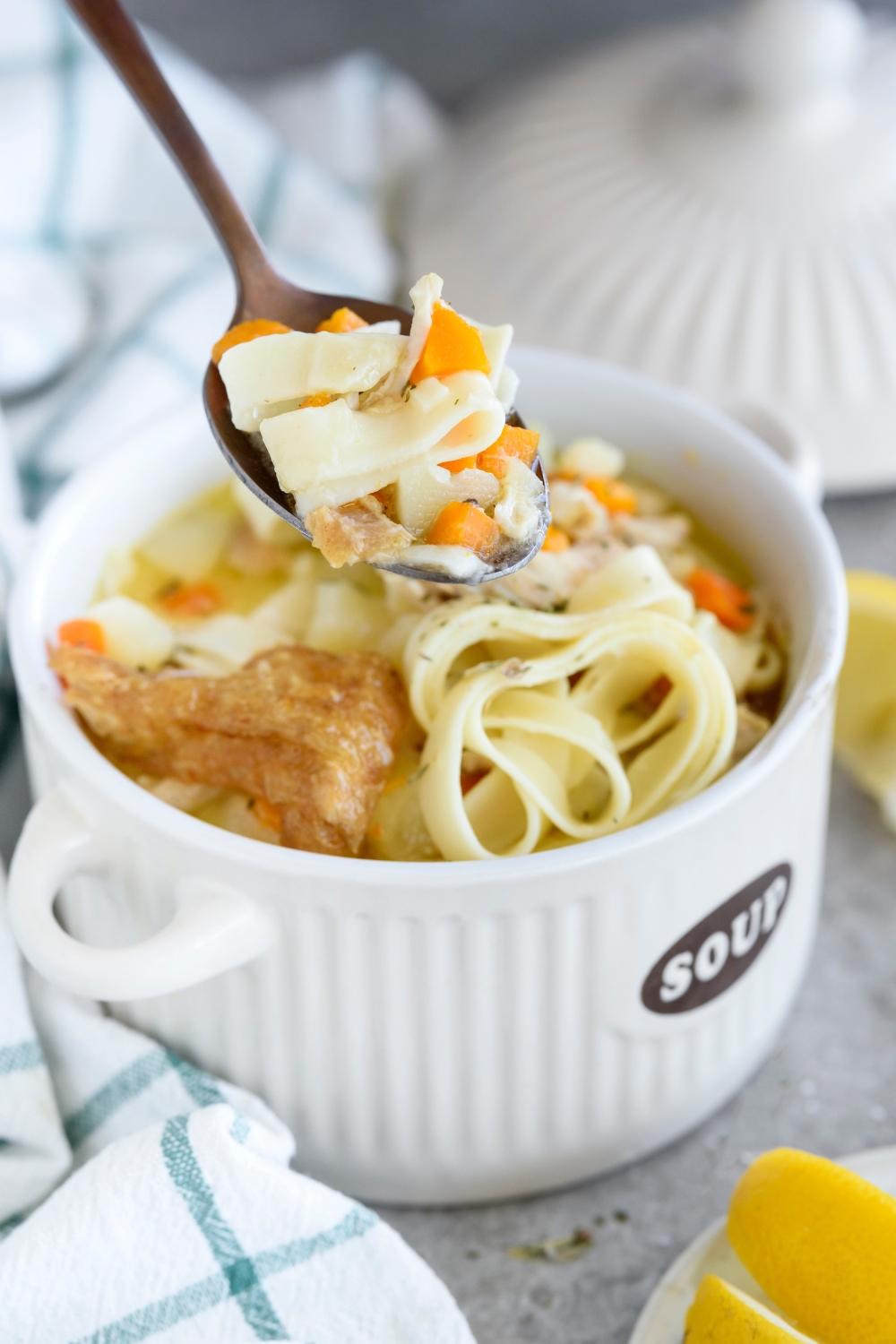 A spoonful of chicken noodle soup held above a bowl of soup, with chunks of carrots and chicken skin.