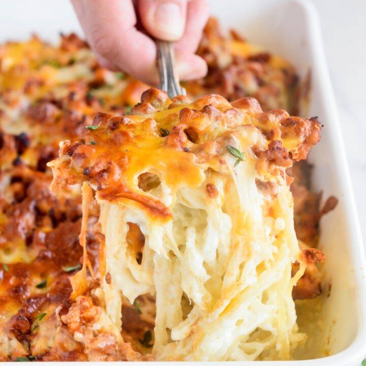 A hand scooping a serving of baked spaghetti from a baking dish. The spaghetti is covered in white sauce and topped with sausage chunks and melted cheese.