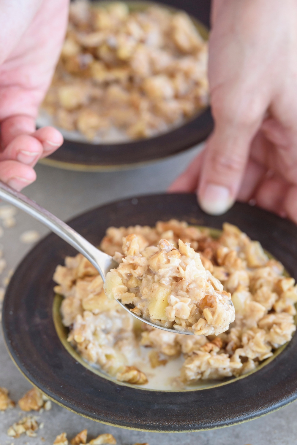A spoonful of oatmeal with diced apples and chopped nuts mixed in. The spoon is held above a bowl filled with oatmeal.