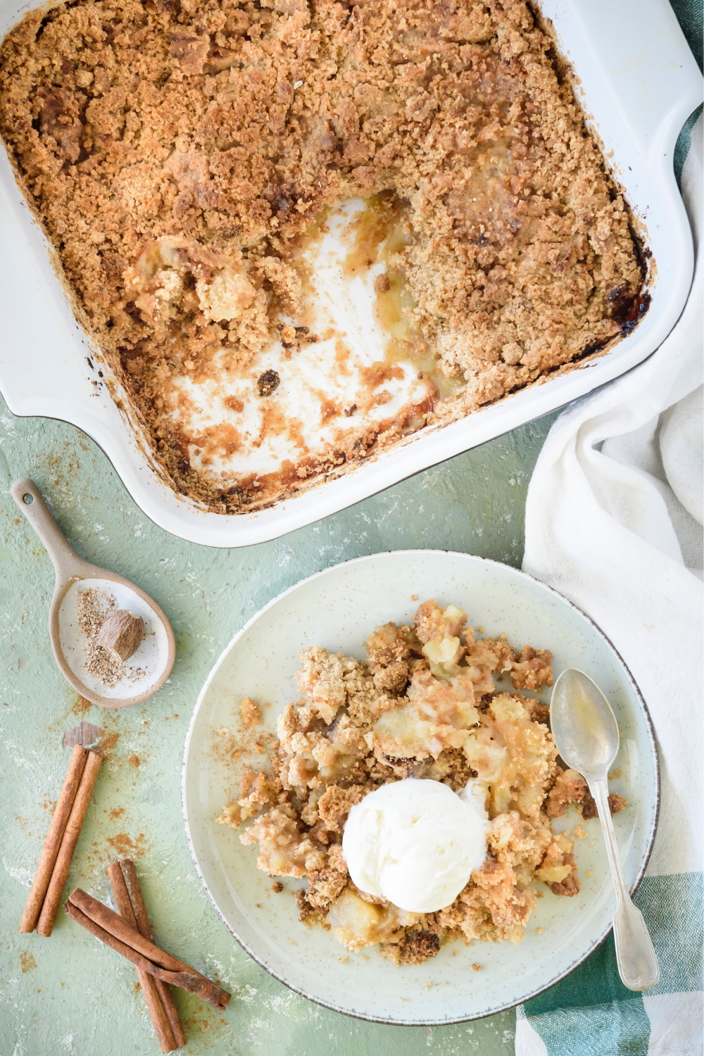 Apple crumble on a plate with a scoop of vanilla ice cream on top and a fork is also on the plate. Next to the plate is a baking dish filled with more apple crumble.