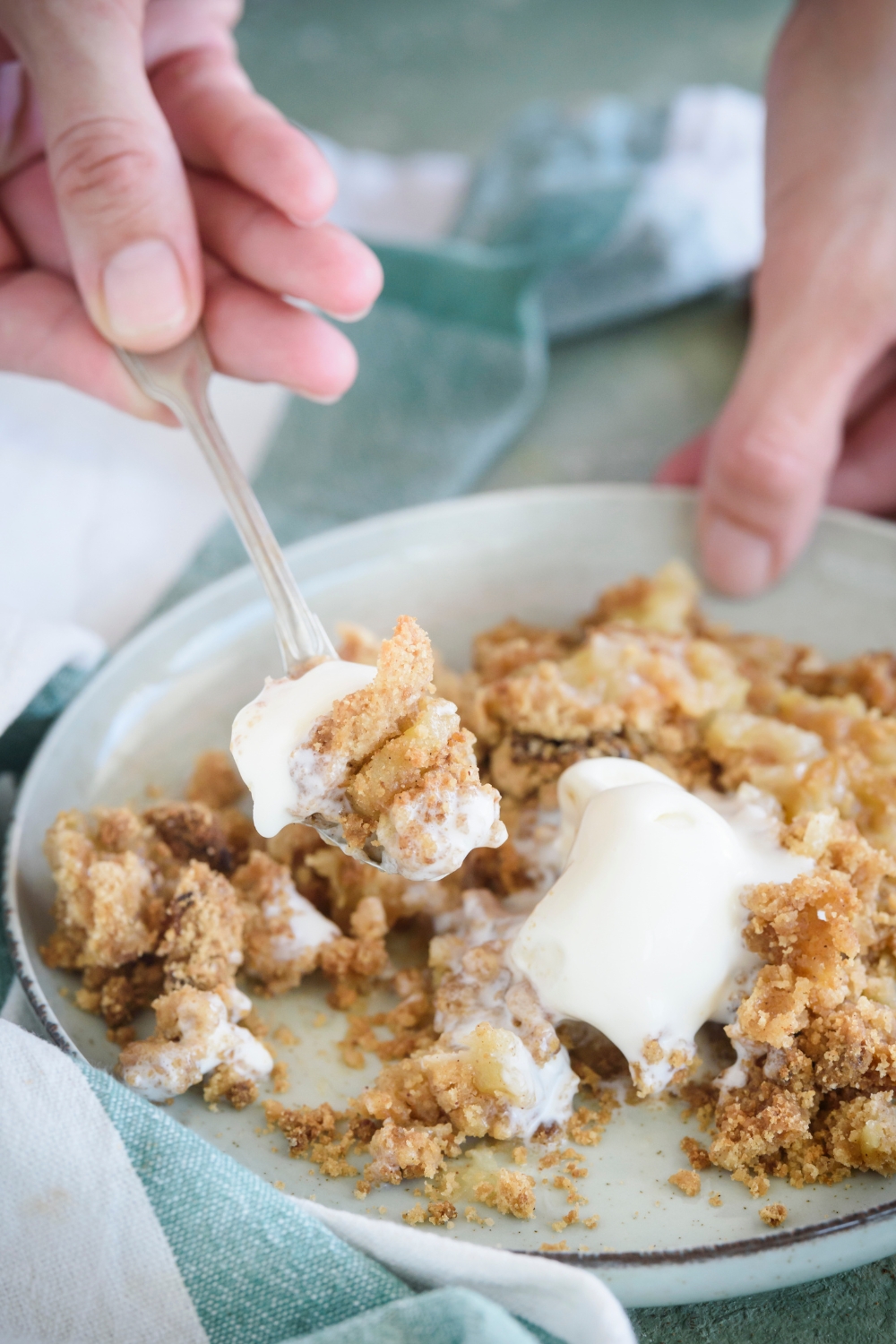 A spoonful of apple crumble with vanilla ice cream dripping off the fork. The spoon is held above the plate filled with more apple crumble.