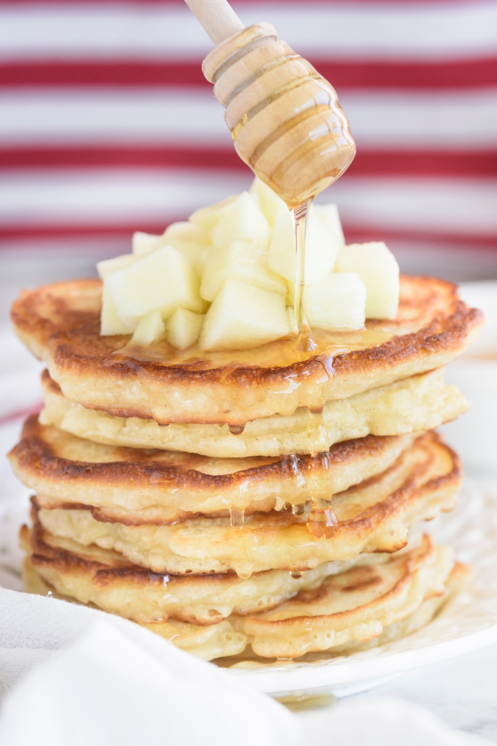 A tall stack of pancakes topped with diced apples and honey being drizzled down the sides of the pancakes.