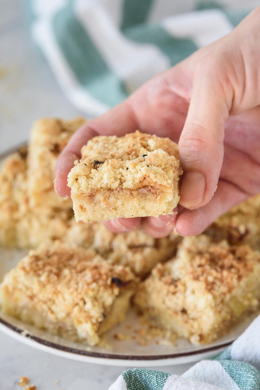 An apple pie bar held above a plate filled with more apple pie bars. The bar is layered with a crust, apple filling, and a crumbly topping.