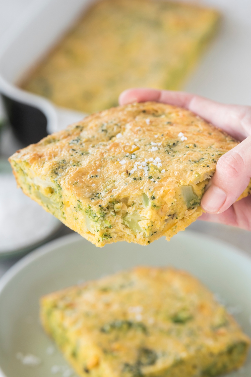 A square of broccoli cornbread garnished with sea salt and a bite has been taken out.