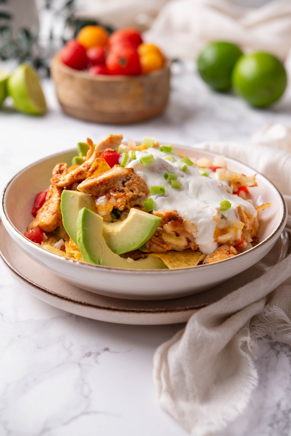 A bowl of chicken nachos covered in queso sauce, garnished with pico de gallo and green onions, with sliced avocado on the side.