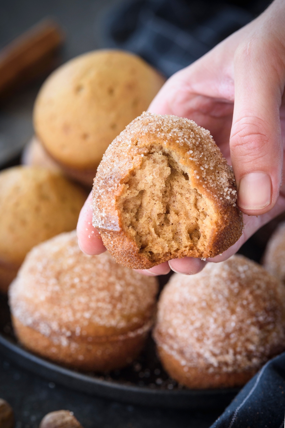 A hand holding a muffin above a plate filled with muffins. A bite has been taken out of the muffin.