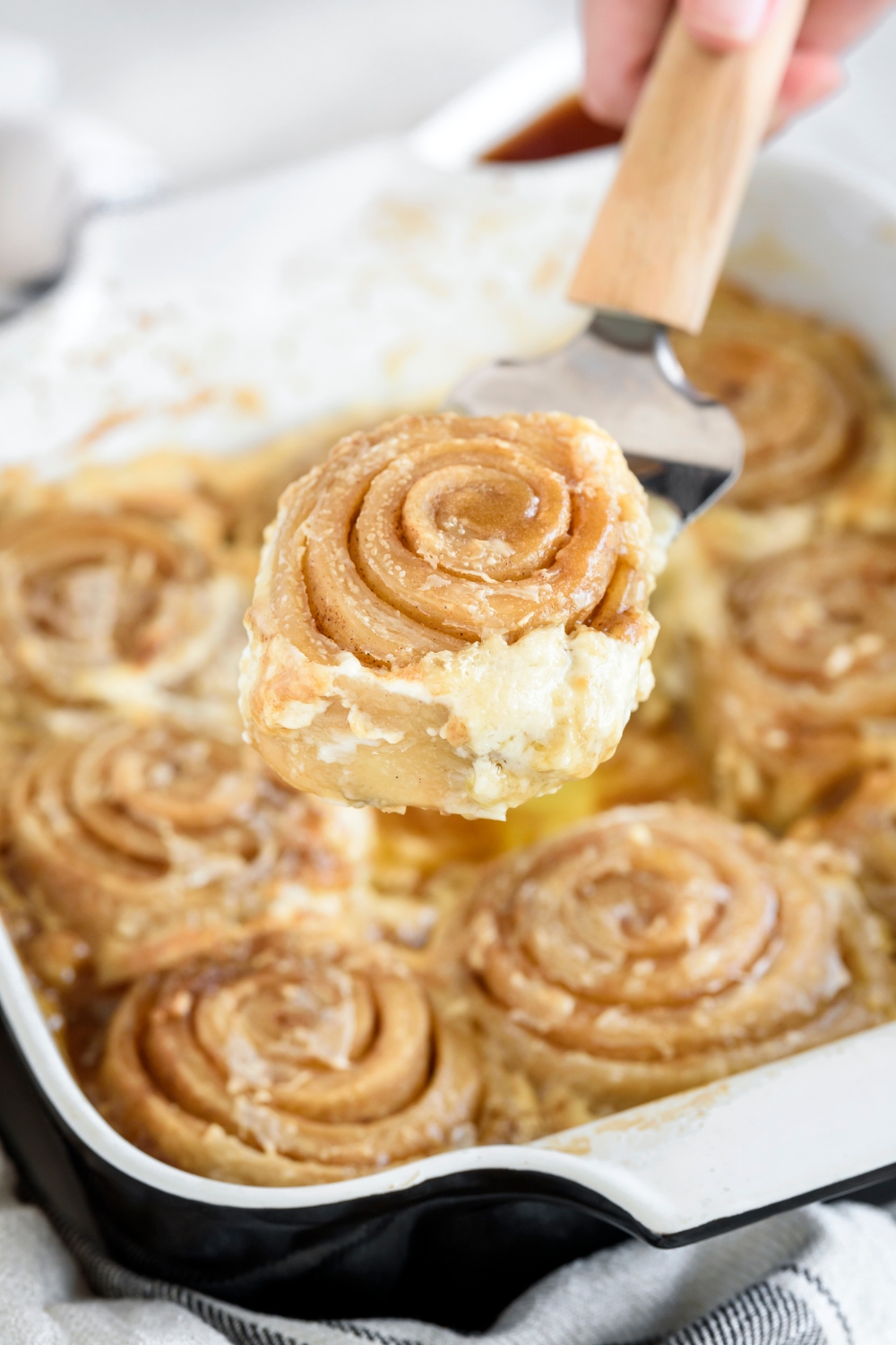 A cinnamon roll on a spatula lifted above a baking dish filled with more cinnamon rolls.