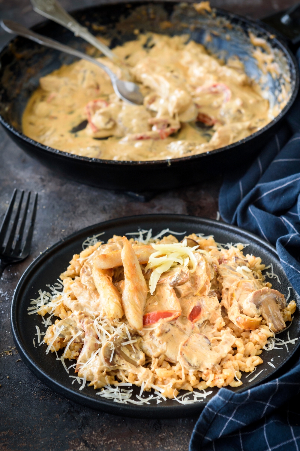 A plate of chicken in cream sauce with red peppers on a bed of rice next to a skillet filled with more chicken and peppers in a seasoned cream sauce.