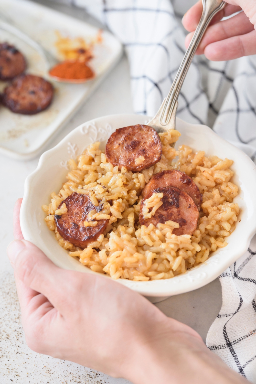 A bowl of dirty rice with four cooked sausage slices on top. There is a fork in the bowl and a person holding the bowl.