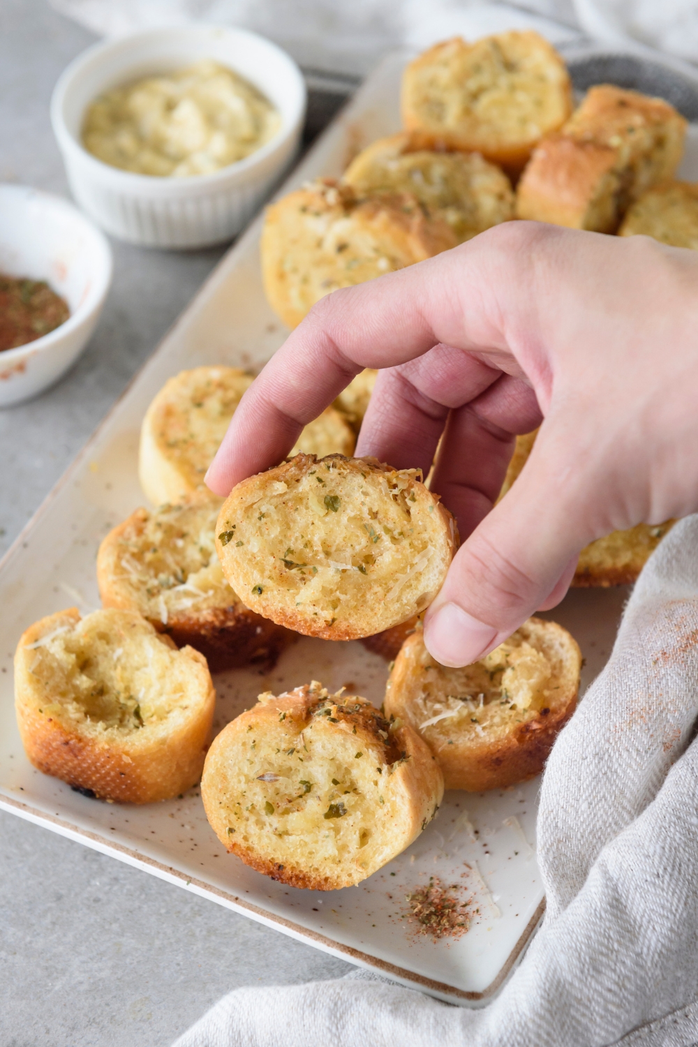 A hand picking up a slice of baguette garlic bread from a plate filled with slices of seasoned garlic bread.