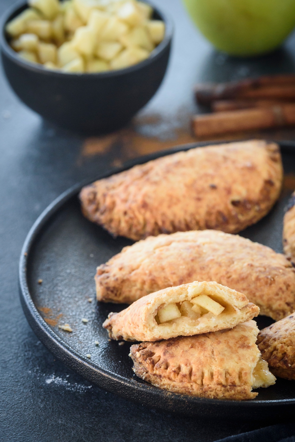 A plate filled with mini apple pies and one has been cut in half with each half stacked on top of each other revealing the apple filling.