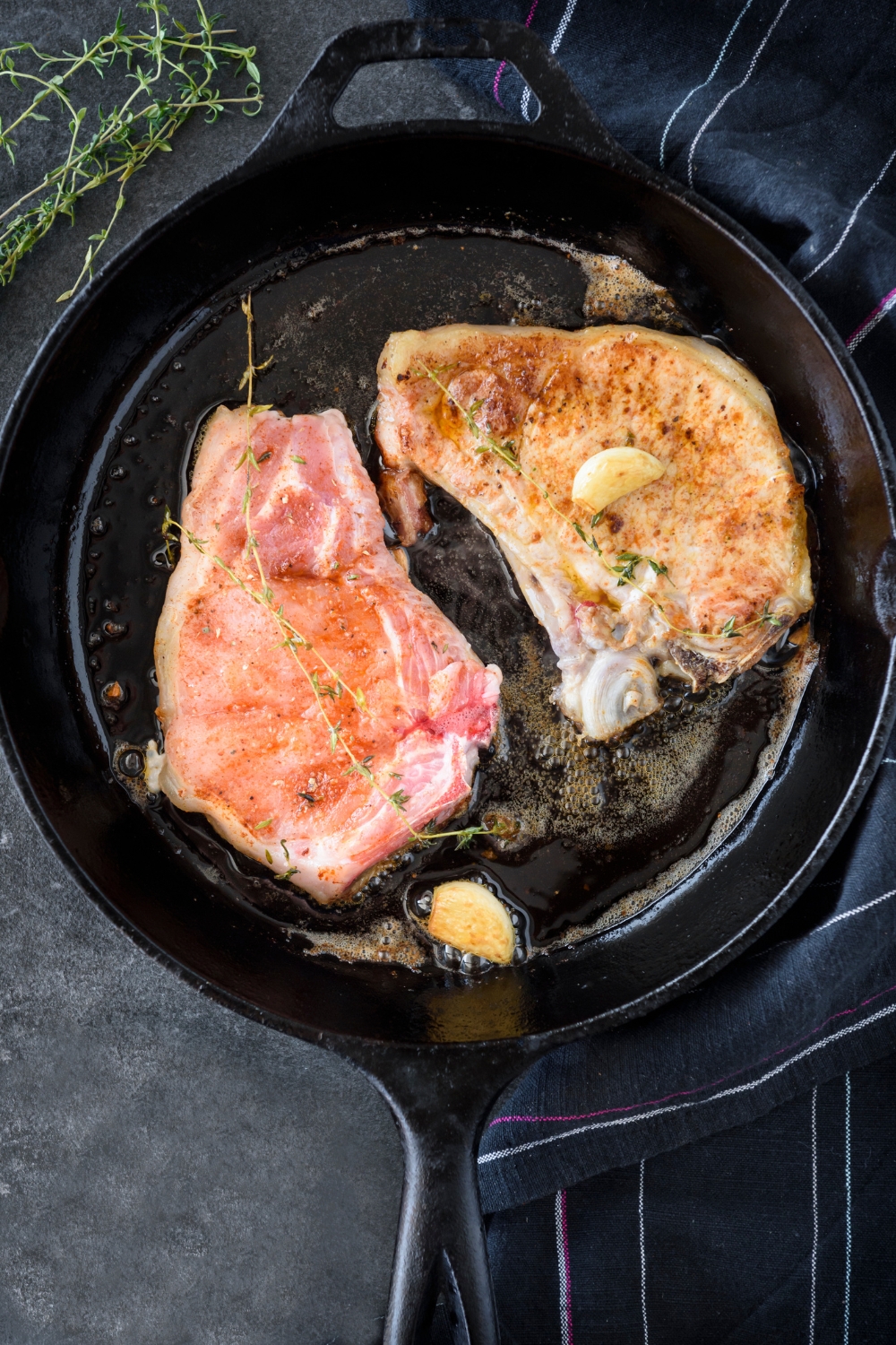 A cast iron skillet with two pork chops cooking in it along with garlic cloves and herbs.