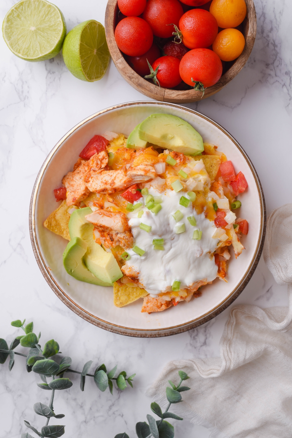 A bowl of chicken nachos covered in queso sauce, garnished with pico de gallo and green onions, with sliced avocado on the side.