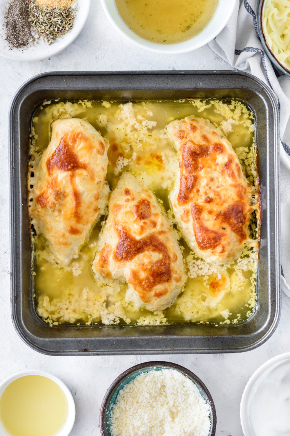 A baking dish filled with three freshly baked chicken breasts. The chicken is golden brown and there is chicken broth partially covering the chicken.