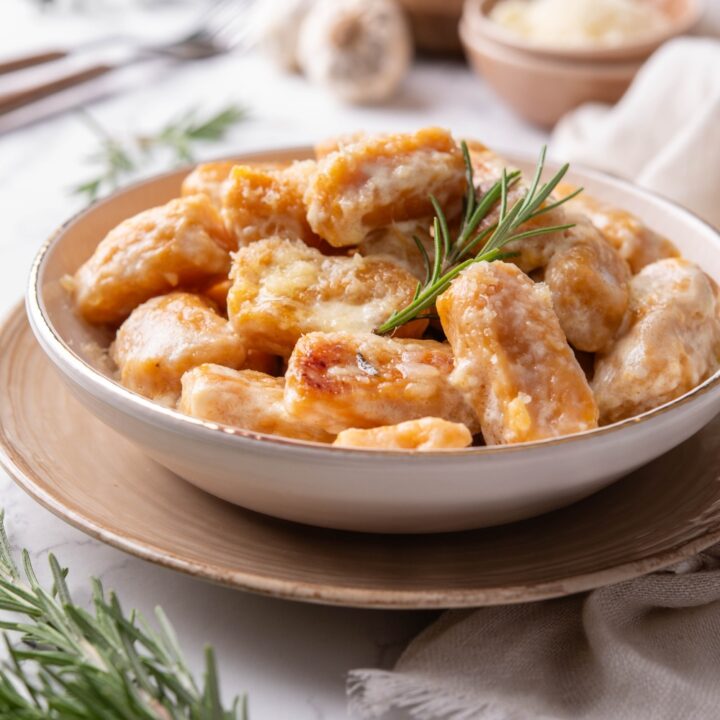 A bowl of sweet potato gnocchi covered in cream sauce with a sprig of rosemary garnished on top.