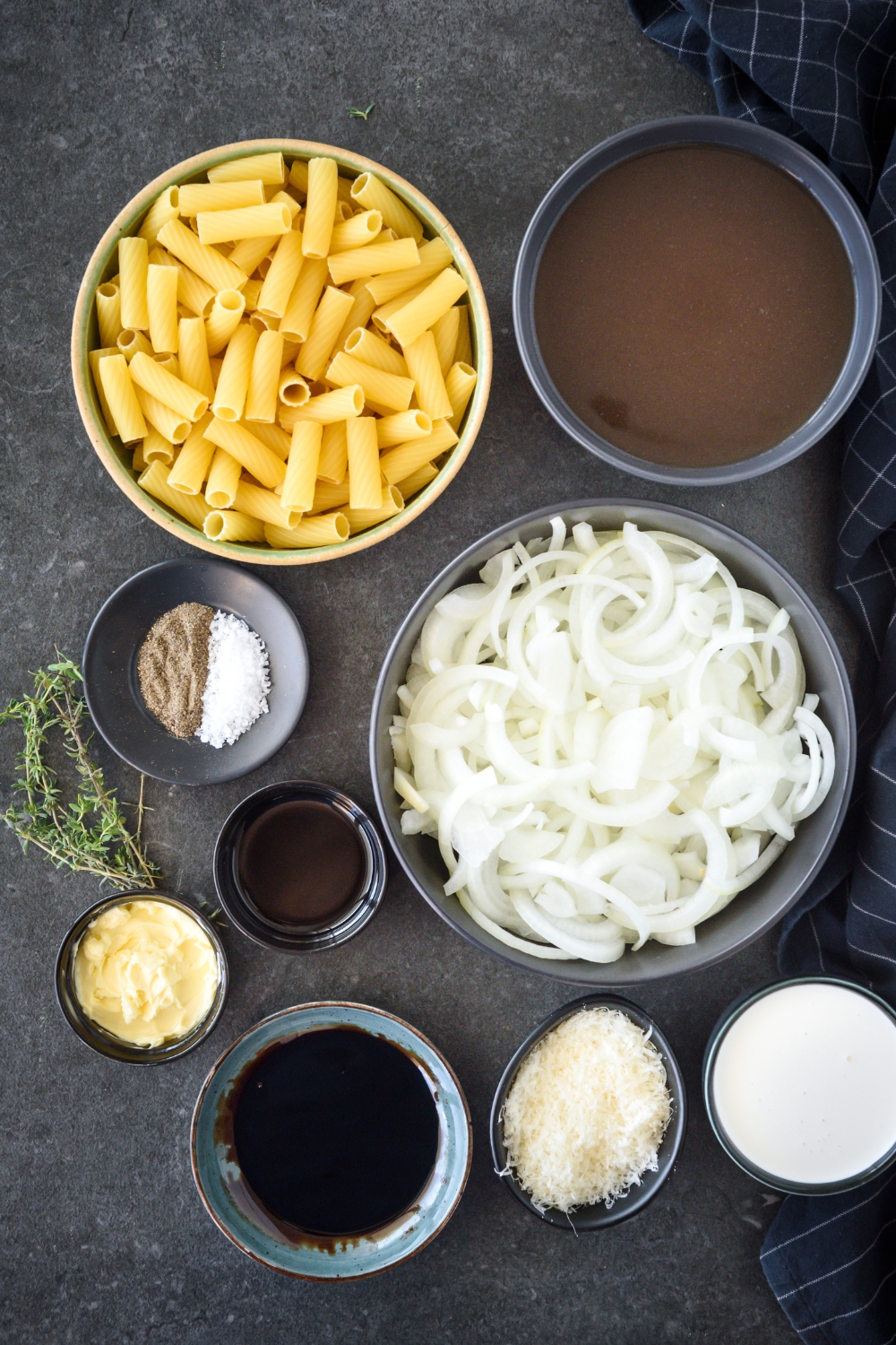 An assortment of ingredients including bowls of dried pasta, broth, sliced onions, cheese, cream, soy sauce, and seasonings on a black counter.