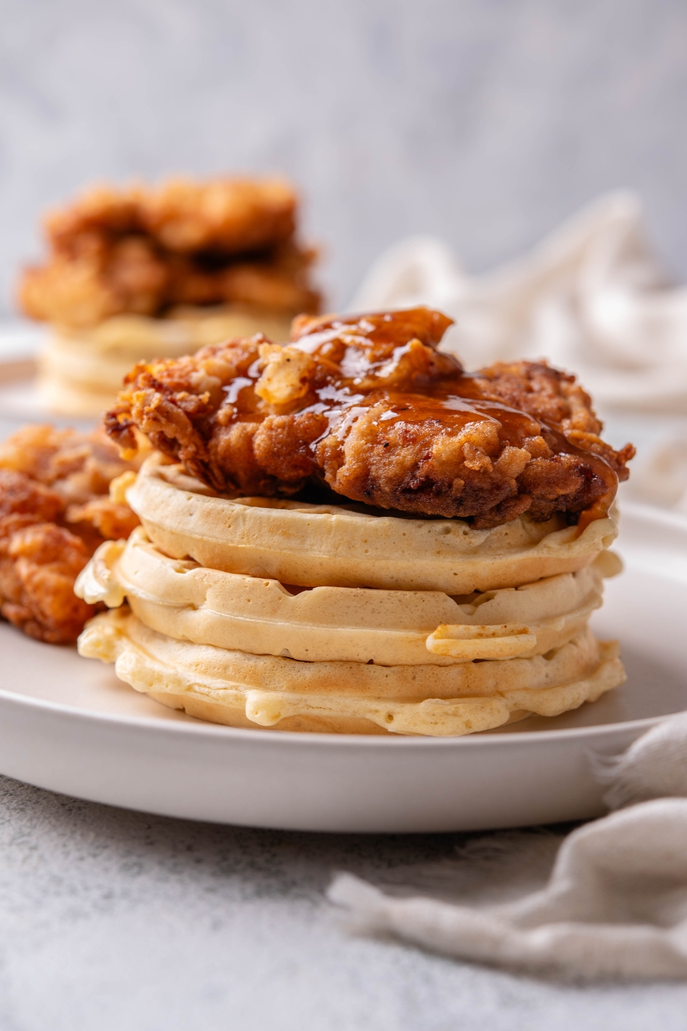 A stack of three waffles with a piece of fried chicken on top covered in a glaze. There is a second piece of fried chicken next to the stack of waffles.