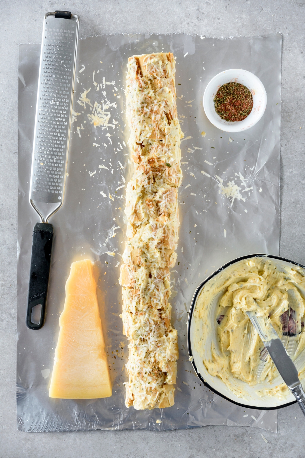 A baguette that's been cut into slices and covered in a seasoned butter mixture. The baguette has been sprinkled with seasonings and is on a sheet of foil next to a wedge of cheese and bowls of seasonings and butter.
