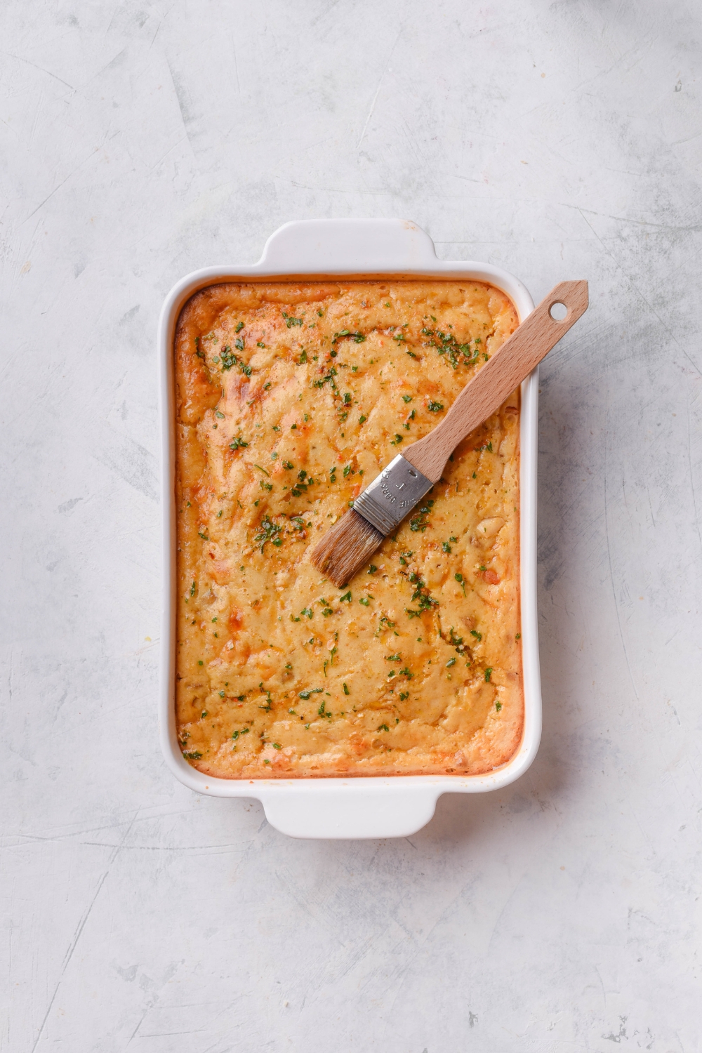 A baking dish filled with freshly baked casserole covered in a biscuit topping that has been brushed with a seasoned butter using a basting brush.
