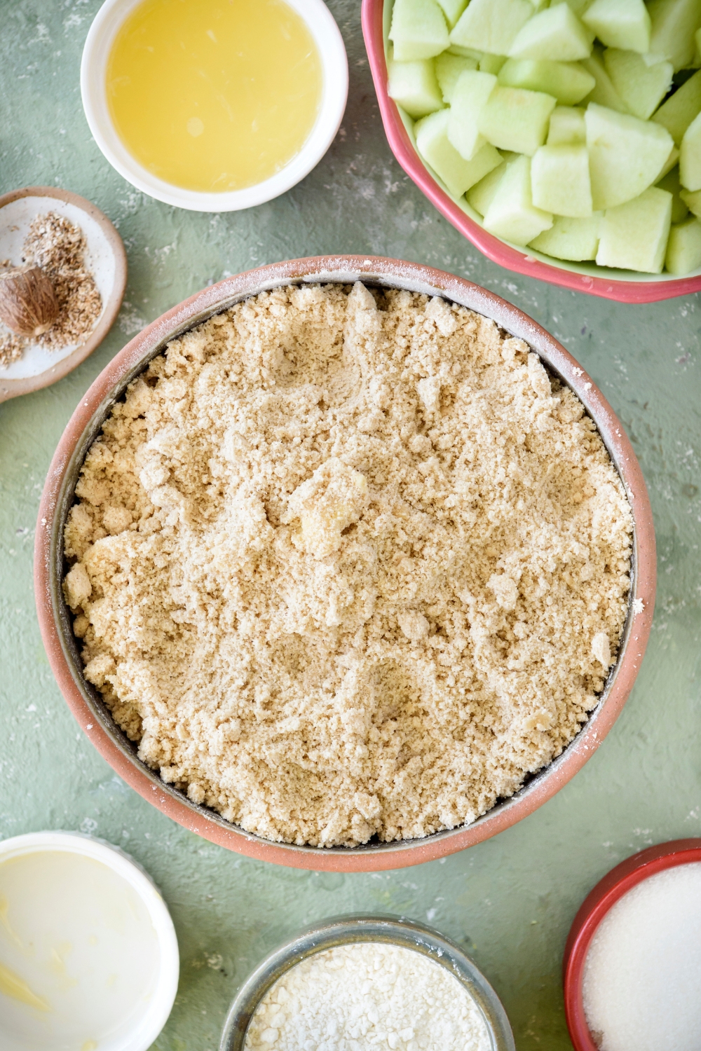 A bowl filled with a crumbly mixture that is surrounded by an assortment of ingredients.